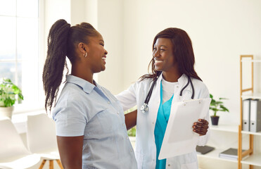 Poster - Portrait of smiling friendly female african american doctor therapist talking with a young woman patient holding report file with appointment standing in office. Medicine and health care concept.