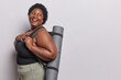 Studio shot of cheerful obese African woman with curly hair laughs positively keeps hand on chest carries rubber fitness mat dressed in active wear goes in for sport to loose weight isolated