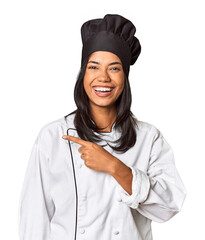 Wall Mural - Young Filipina chef with cooking hat in studio smiling and pointing aside, showing something at blank space.