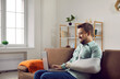 © Studio Romantic - Smiling man with fractured arm sitting on sofa and using laptop computer. Handsome man with plastered arm in sling bandage sitting on couch working or chatting online while recovering at home