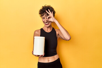 Wall Mural - MIddle aged athlete woman holding protein supplement on yellow excited keeping ok gesture on eye.