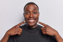 Young positive dark skinned man points at toothy smile shows perfect teeth dressed in casual black t shirt attracts your attention to mouth dressed in casual black t shirt isolated on white background