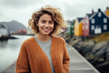 Wall Mural - Portrait of a grinning woman in her 20s wearing a chic cardigan against a picturesque seaside village. AI Generation