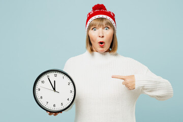 Merry shocked elderly woman 50s years old wear sweater red hat posing hold in hands point index finger on clock isolated on plain blue background. Happy New Year celebration Christmas holiday concept.