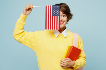 Wall Mural - Young fun woman student wear casual clothes yellow sweater backpack bag hold cover half of face with notebook American flag isolated on plain blue background. High school university college concept.