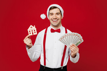 Wall Mural - Merry young fun man wear white shirt Santa hat posing hold fan cash money in dollar banknotes wooden house mockup isolated on plain red background Happy New Year Christmas celebration holiday concept