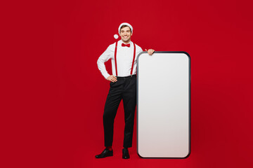 Wall Mural - Full body merry young fun man wear white shirt Santa hat posing big huge blank screen mobile cell phone isolated on plain red background studio. Happy New Year Christmas celebration holiday concept.