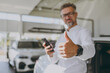 © ViDi Studio - Adult man customer male buyer client wear shirt use mobile cell phone choose auto want buy new automobile in car showroom vehicle salon dealership store motor show. Sales concept. Focus on thumb up.