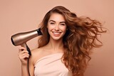 A smiling woman dries her hair with a hair dryer, on a beige background.