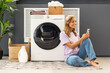 Young attractive cute woman, housekeeper holding smartphone using mobile app sitting on carpet in modern laundry room with washing machine, laundry detergent, basket with clothes on background. Mockup