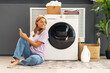 Beautiful smiling woman, housewife holding mobile phone sitting on carpet looking away in modern laundry room with washing machine, laundry detergent, basket with clothes on background. Advertisement