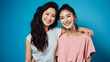 Two Asian girls smiling and hugging on blue background. Best friends or female partner, free and inclusive friendship and love.