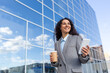 canvas print picture - Successful Hispanic businesswoman walks to the office, holds a coffee and a phone in her hands, smiles and solves cases on the way to work, stands on the street of the office.