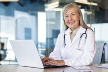 Portrait Of Mature Experienced Female Doctor, Gray-haired Senior Woman In White Medical Coat Smiling Looking At Camera, Consulting Patients Remotely, Sitting At Table With Laptop Inside Clinic.