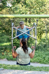 Son and mother swing hard on a double swing in a green summer apple orchard