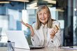 Portrait of a senior gray-haired business woman looking worriedly at the camera, sitting in the office at the table with a laptop and holding a credit card in her hand.