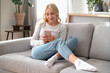 © brizmaker - Dreamy woman sitting on couch, using modern smartphone and smiling
