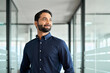 Happy bearded Indian business man leader looking away standing in office hallway. Professional smiling businessman manager executive or male employee or entrepreneur thinking of future success.