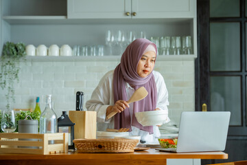 Wall Mural - muslim mother looking at recipe from laptop and cooking. having fun woman with hijab preparing dinner, Islamic woman Enjoying Doing Homemade, Teaching cooking online