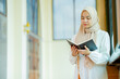 © maya1313 - The image of an Asian Muslim woman in the Islamic religion in hijab in cream color. reading the Quran and having a happy Staying in a beautiful mosque, Arabic word translation: The Holy Al Quran.