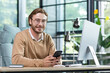 © Liubomir - Friendly businessman in glasses is working in the office at the table, holding the phone in his hands, texting, smiling and looking at the camera.