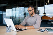 © Liubomir - Serious African American businessman working in modern office, sitting at desk in front of laptop, tapping on keyboard, modern work concept.