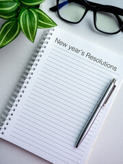 Wall Mural - Goals, action, plan, business direction and to do list concepts in new year. Text, New year's Resolutions on blank notebook page with pen near glasses and green plant on vertical white background.