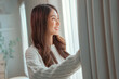 Close up of happy young asian woman opening curtain at home, Smiling attractive lady looking outside apartment enjoying dreaming and contemplating feeling hope concept