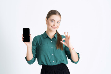 Attractive young woman shows an empty mobile phone display with a copy space for advertising and shows an OK gesture isolated on a white studio background. Advertising of the application