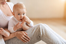 Adorable Infant Baby Boy Sitting In Mother Arms, Chewing Feet