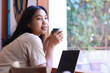 © M Alfan Setyawan - happy asian woman sitting in cafe drinkking coffee and working with laptop near window wearing fashionable clothes, contemplative female remote worker holding mug in coffee shop look away