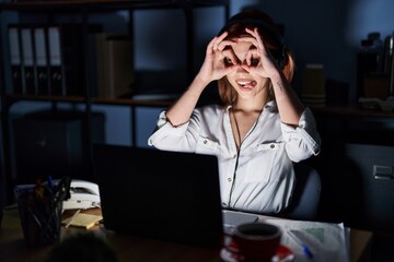 Wall Mural - Young caucasian woman working at the office at night doing ok gesture like binoculars sticking tongue out, eyes looking through fingers. crazy expression.