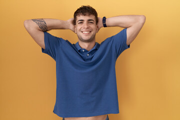 Poster - Young hispanic man standing over yellow background relaxing and stretching, arms and hands behind head and neck smiling happy