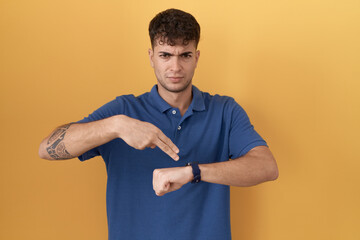 Poster - Young hispanic man standing over yellow background in hurry pointing to watch time, impatience, upset and angry for deadline delay