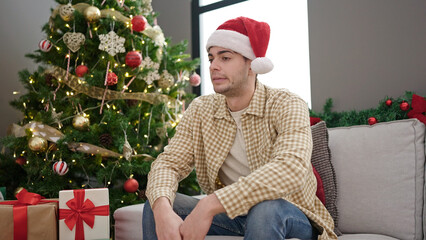 Poster - Young hispanic man sitting on sofa by christmas tree looking upset at home