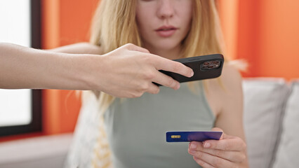 Poster - Young blonde woman make photo to credit card by smartphone sitting on sofa at home