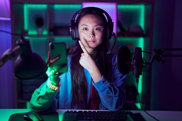 Poster - Young asian woman playing video games with smartphone pointing with hand finger to the side showing advertisement, serious and calm face