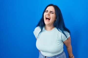 Canvas Print - Young modern girl with blue hair standing over blue background angry and mad screaming frustrated and furious, shouting with anger. rage and aggressive concept.