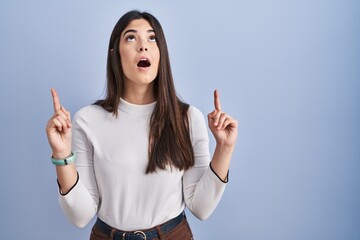 Wall Mural - Young brunette woman standing over blue background amazed and surprised looking up and pointing with fingers and raised arms.