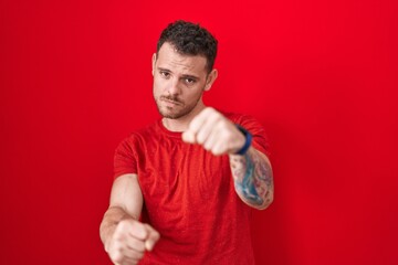 Poster - Young hispanic man standing over red background punching fist to fight, aggressive and angry attack, threat and violence