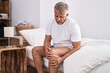 Middle age grey-haired man suffering for knee injury sitting on bed at bedroom