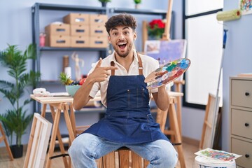 Arab man with beard painter sitting at art studio holding palette smiling happy pointing with hand and finger