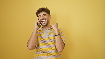 Canvas Print - Upbeat young arabian man celebrating his win, confidently talking on smartphone, standing with a big smile against a yellow isolated background