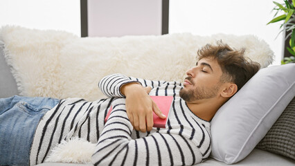 Wall Mural - Exhausted young arab man finding comfort, sleeping peacefully on his cozy sofa at home, book clutched in hands after reading