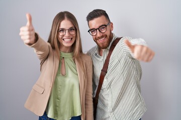 Wall Mural - Young couple standing over white background approving doing positive gesture with hand, thumbs up smiling and happy for success. winner gesture.