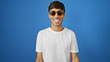 Cheerful young hispanic man wearing sunglasses, exuding confidence and positivity, standing against an isolated blue background while enjoying a cool, casual lifestyle.
