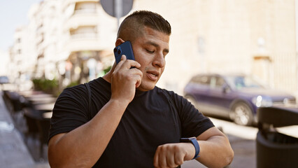 Canvas Print - Casual latin man, talking seriously on his smartphone, checks the time on his wristwatch while standing under the sunny urban city outdoors.