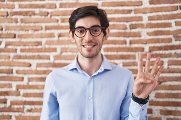 Wall Mural - Young hispanic man standing over brick wall background showing and pointing up with fingers number four while smiling confident and happy.