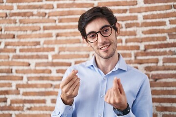 Wall Mural - Young hispanic man standing over brick wall background doing money gesture with hands, asking for salary payment, millionaire business