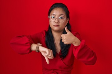 Poster - Asian young woman standing over red background doing thumbs up and down, disagreement and agreement expression. crazy conflict